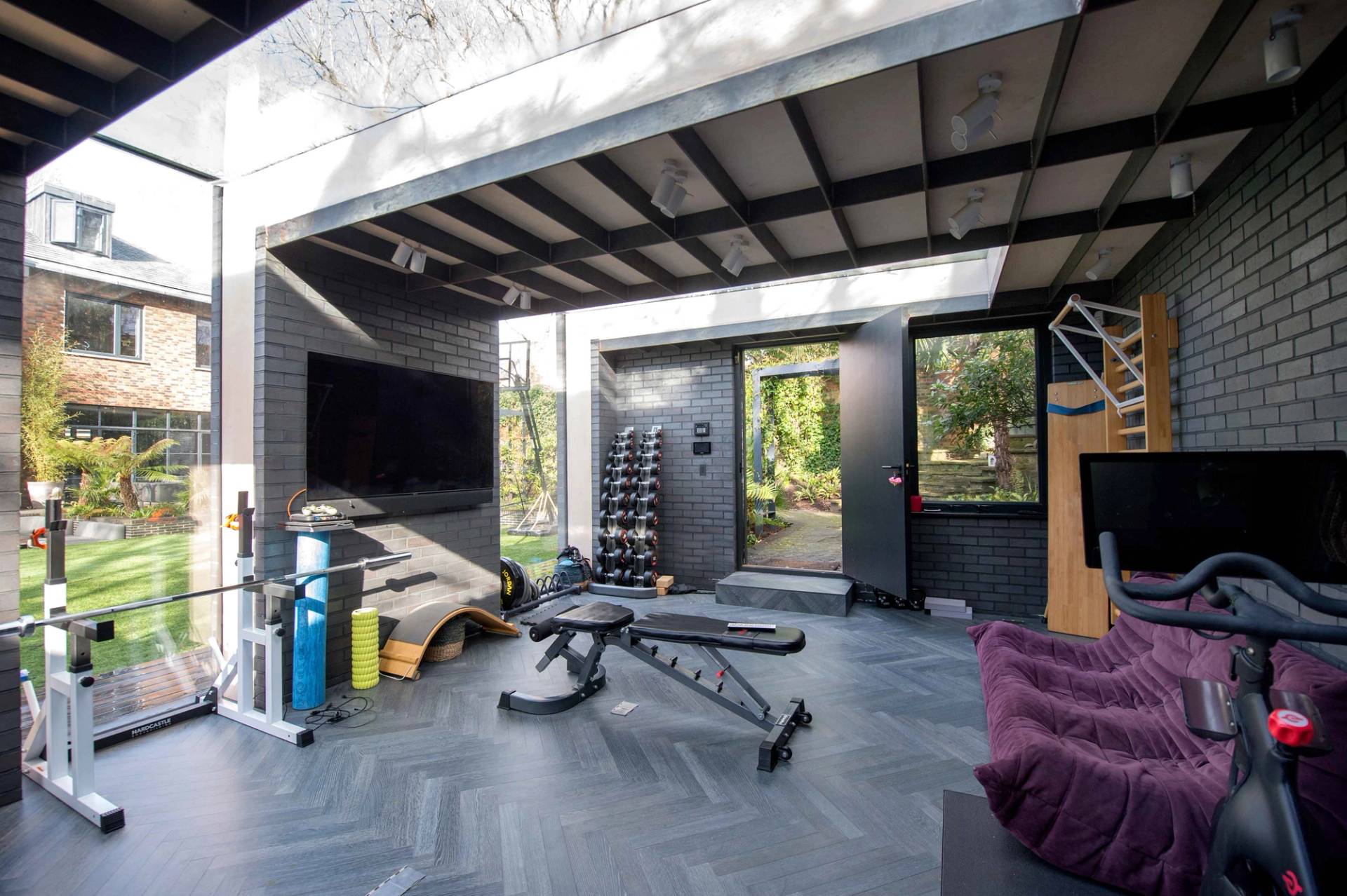 Fully equipped home gym studio featuring heavy-duty fitness gear, rubber flooring, and garden views through massive glass panels, Love Locations.