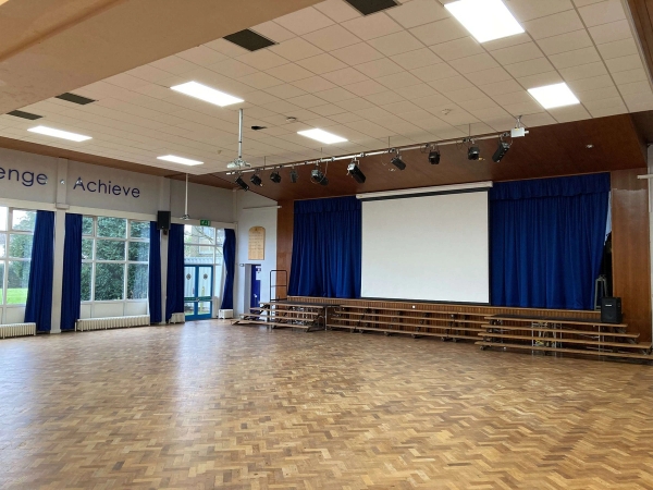 Love Locations modern secondary school hall shoot location Chelmsford Essex CM1: Large assembly hall featuring a wooden parquet floor, theatrical stage, blue curtains, and projector screen, ideal for school plays, TV dramas, and film productions.