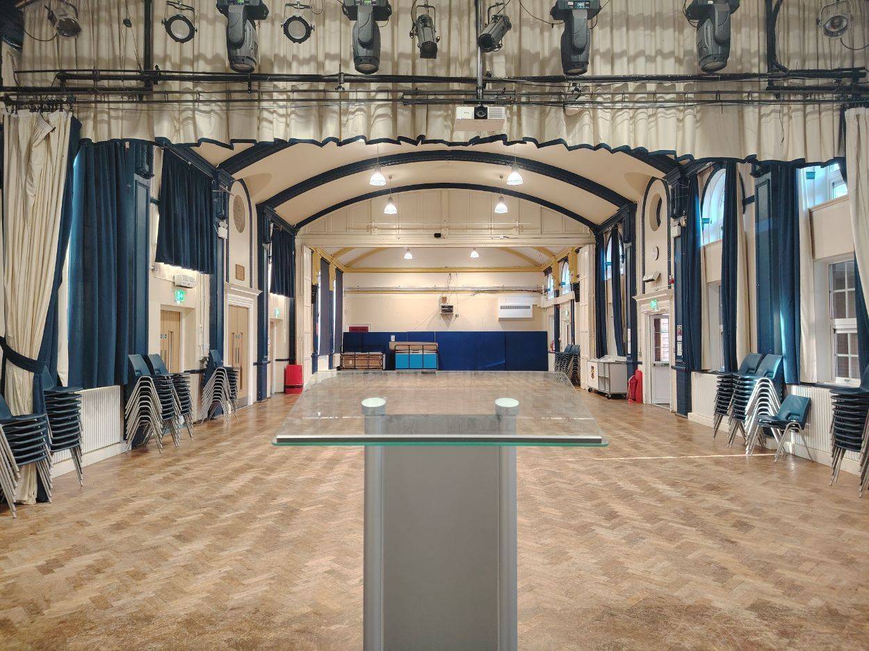 Traditional grammar school assembly hall film location in Sittingbourne Kent ME10 from Love Locations, featuring parquet wood flooring, arched ceilings, blue drapes, and a central podium.