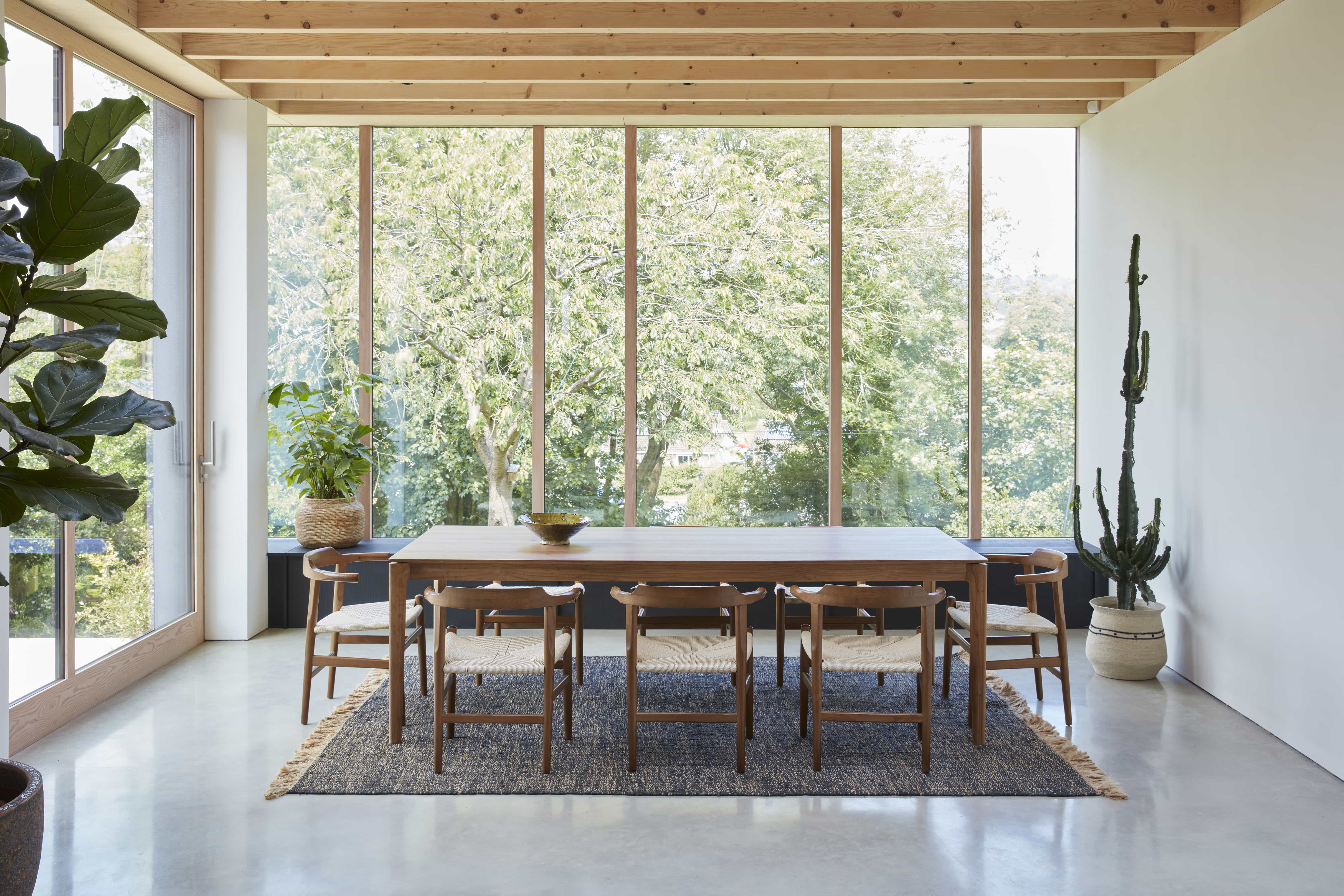 Crescent House – Love Locations. Neutral modern rustic dining area featuring exposed rafter beams, wooden and concrete flooring, floor-to-ceiling windows, a long wooden dining table, 21st-century modern light bentwood chairs, and a cactus in the corner.