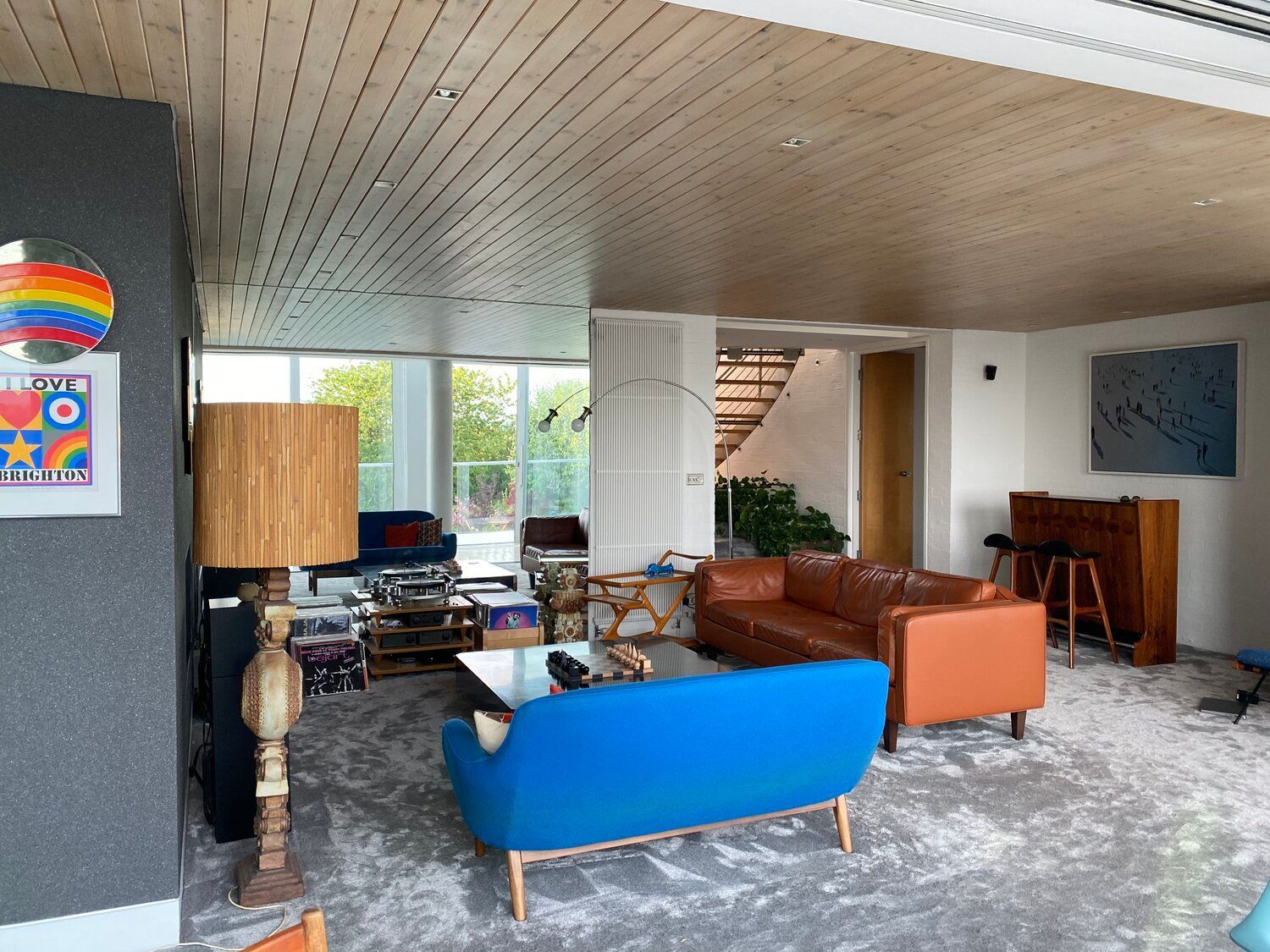 Retro-style room with wooden tongue-and-groove ceiling, blue sofa with angular metal legs, vintage brown leather sofa, grey feature wall, bright 1960s-style artwork, raffia standing lamp, grey carpet, open staircase, large picture windows, and a piano.