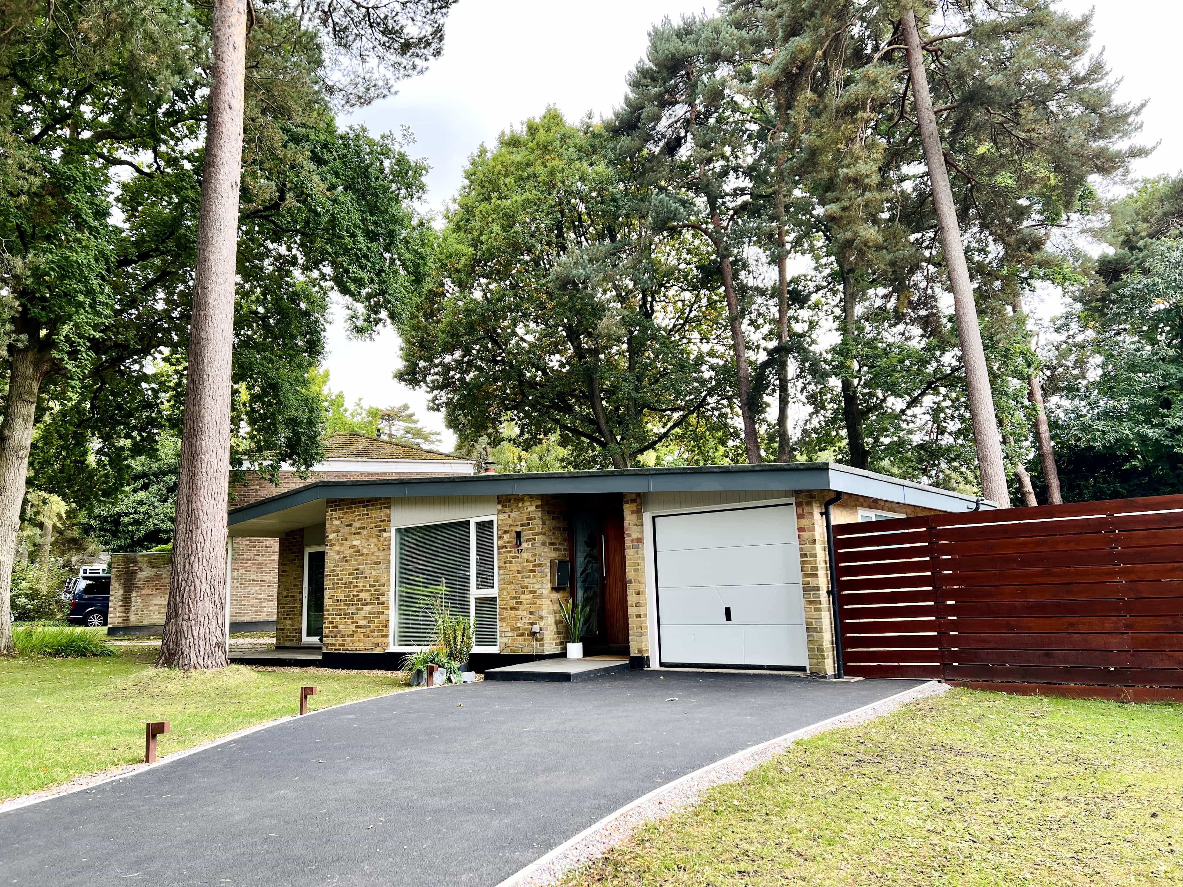 21st-century American-style suburban bungalow with yellow brick exterior, large picture windows, garage door, manicured lawn, driveway, and surrounding mature trees.