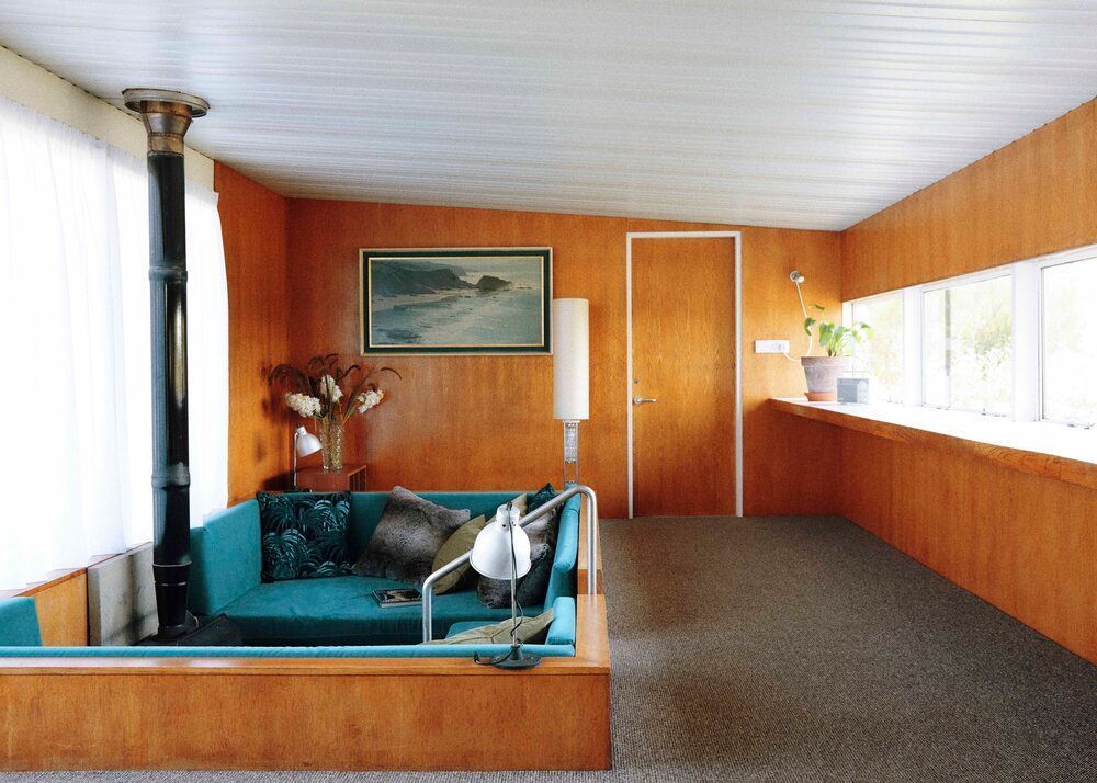 21st-century retro interior with wooden panelled walls, built-in sky blue seating area, black stove, and large surrounding windows.