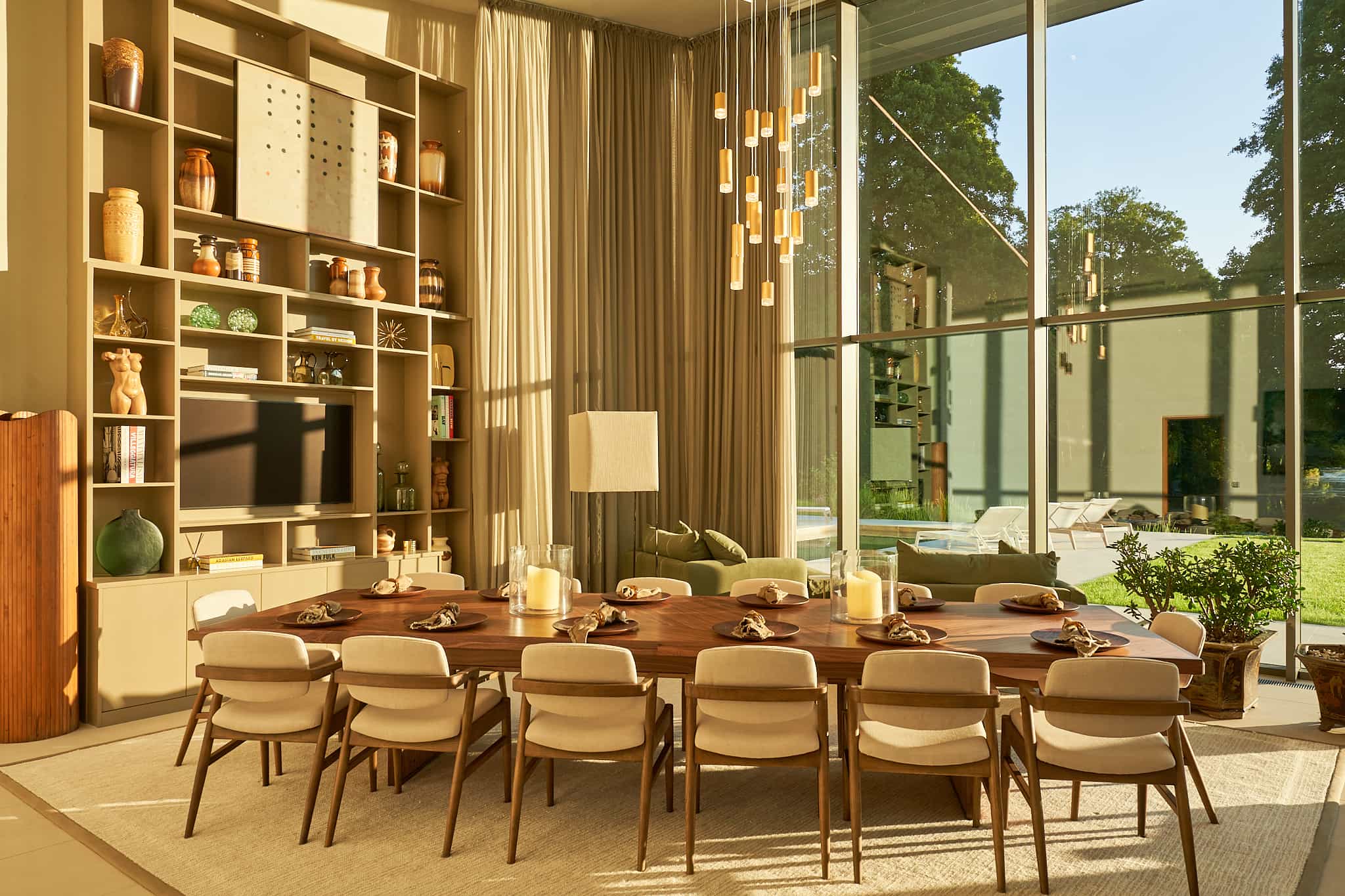 Winchester – Love Locations. High-spec luxury space with a neutral palette, cream drapes, huge windows overlooking a Hollywood-style pool, pale wood shelves with global decor, and a modern dining area with gold chandeliers.