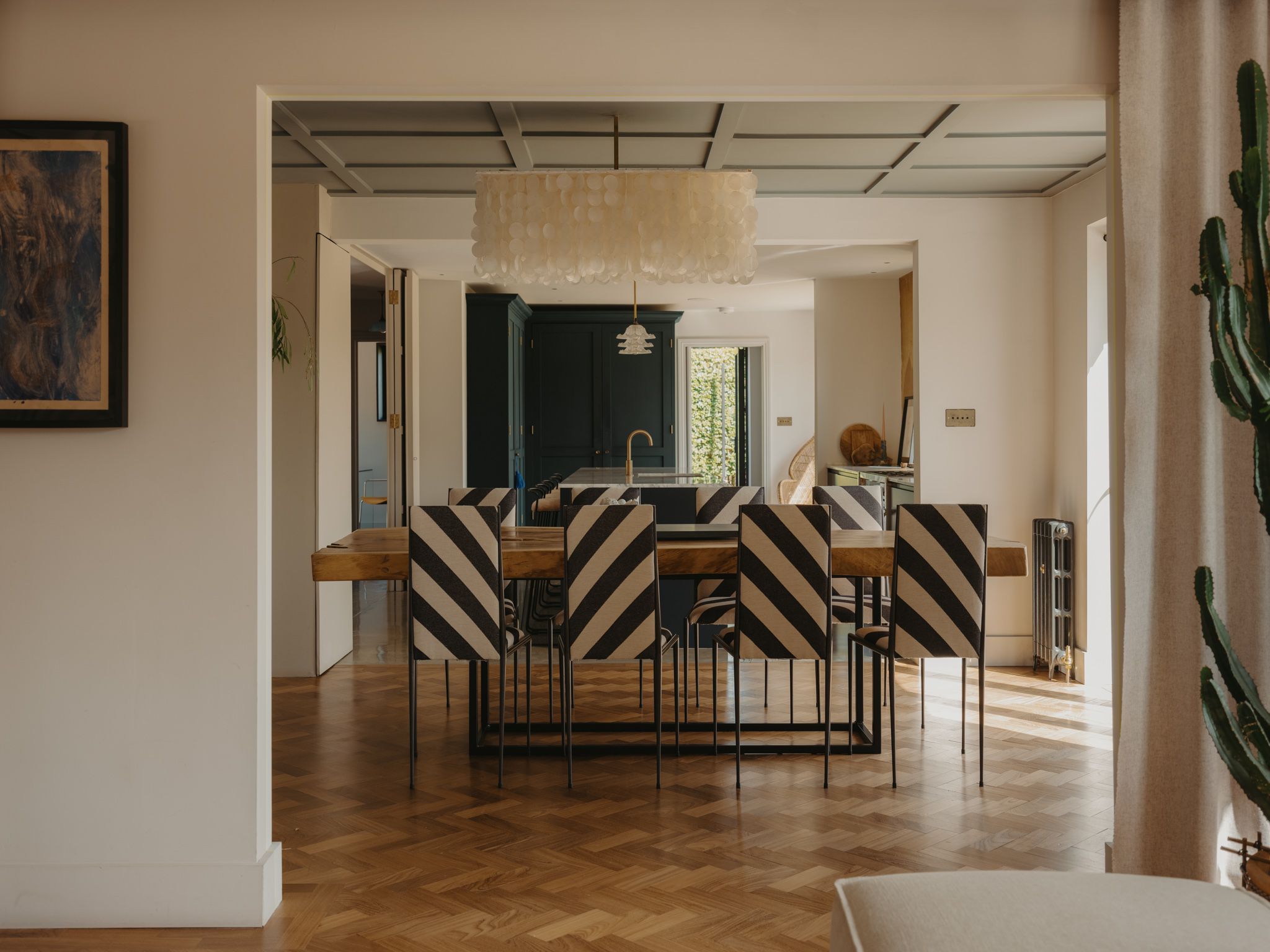 The Hatch – Love Locations. Neutral dining space with modern parquet wooden floor, rustic table, black and cream upholstered metal-legged chairs, dark accent wall, and a striking shell feature chandelier