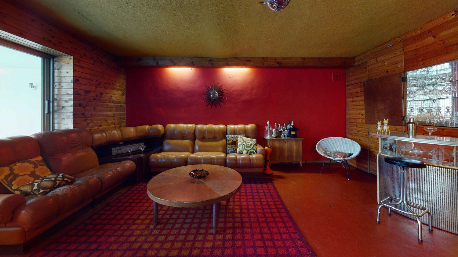 Midcentury retro-style living room with low light brown leather furniture, deep orangery-red walls, orange flooring, red check rug, and a silver and glass retro bar with matching stools.