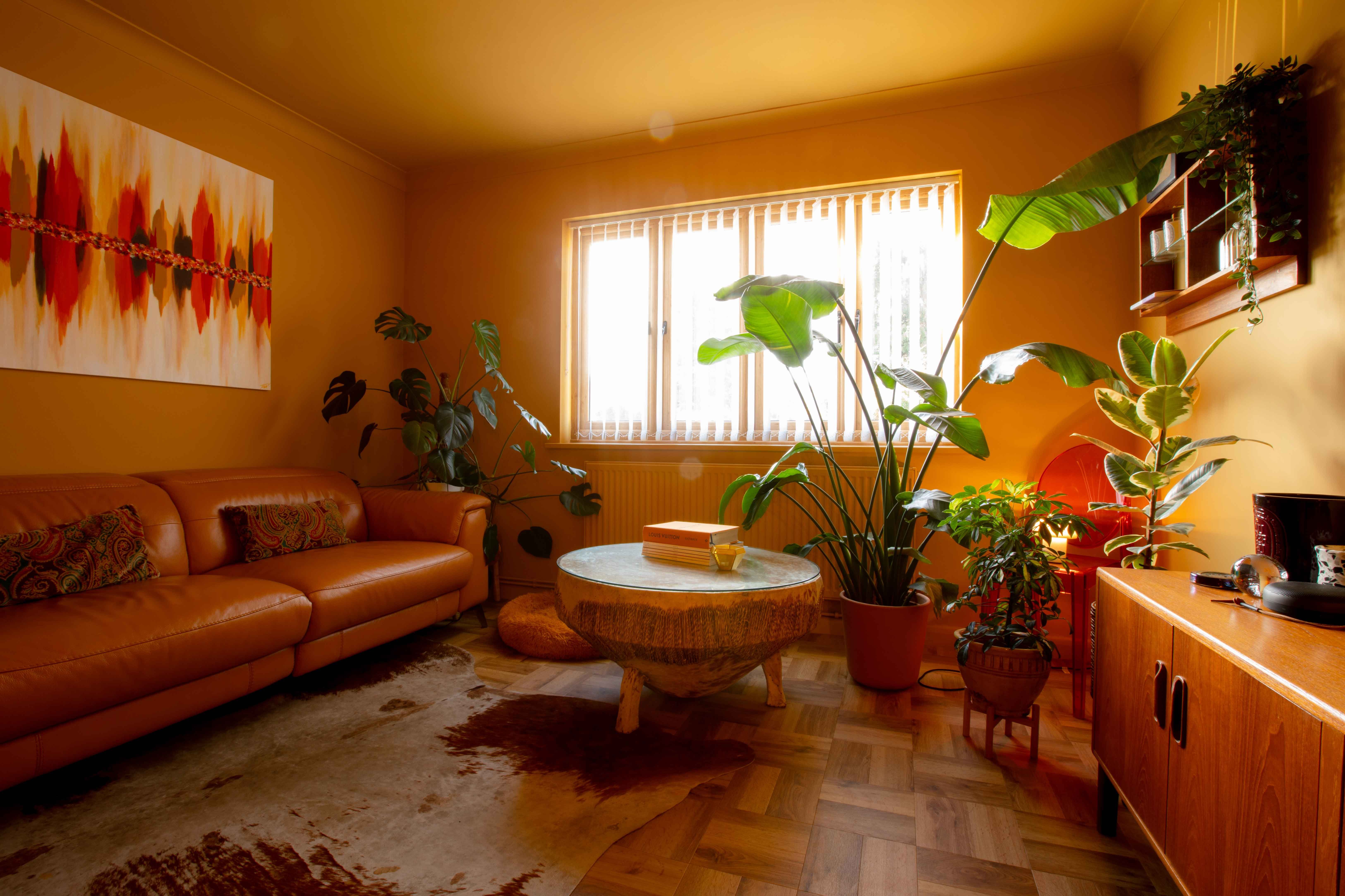 Retro-style room painted in yellow-orange tones with low-level vintage sofa, 60s sideboard, midcentury coffee table, parquet flooring, abstract red-toned painting on the wall, and vertical white blinds on the window—cool, nostalgic vibes.