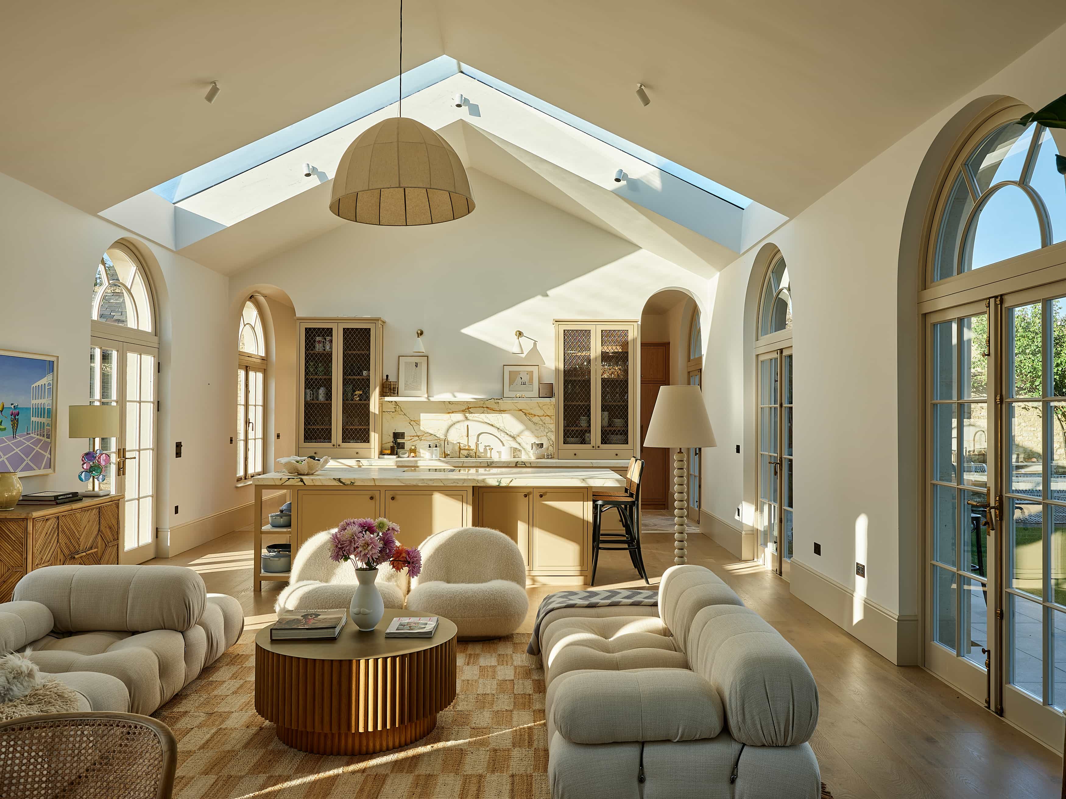 Cotswolds Farm – Love Locations. Modern rustic open-plan kitchen with cream-painted cupboards and a large cream island topped with marble. Sitting room in the foreground features a cream low-lying seating area, wooden textured sideboard, cane armchair, round reeded wooden coffee table, and large arched windows, all evoking a Soho Home style vibe.