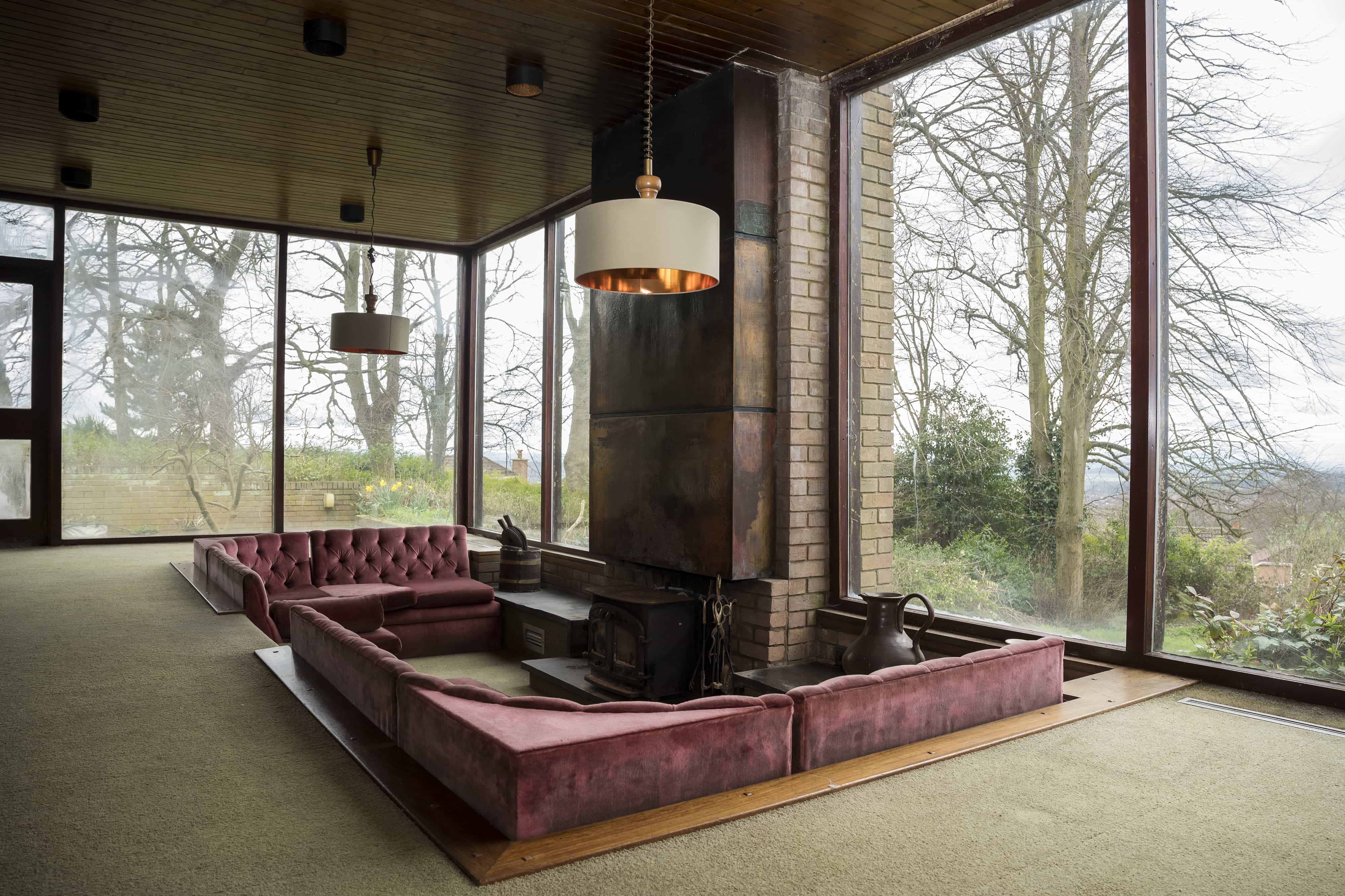 Sunken 1960s-style sofa in an open-plan space with exposed brickwork, floor-to-ceiling windows overlooking a country view, copper-surround fireplace, khaki carpeted flooring throughout, and low-hanging cream and gold interior pendant ceiling lights—groovy vintage aesthetic.