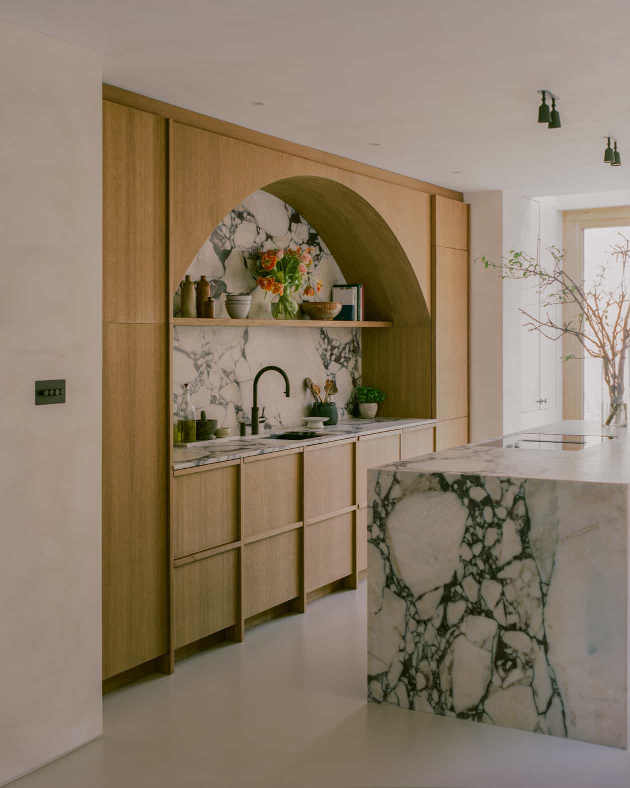 Abbeville Home – Love Locations. Neutral kitchen featuring a wooden arch with a marble backsplash, wooden flat-framed cupboards, and a marble island.
