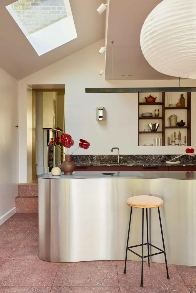 Contemporary stainless steel kitchen with a curved metal island, terrazzo flooring, open shelving, sculptural lighting, and warm neutral tones at Lyon location.