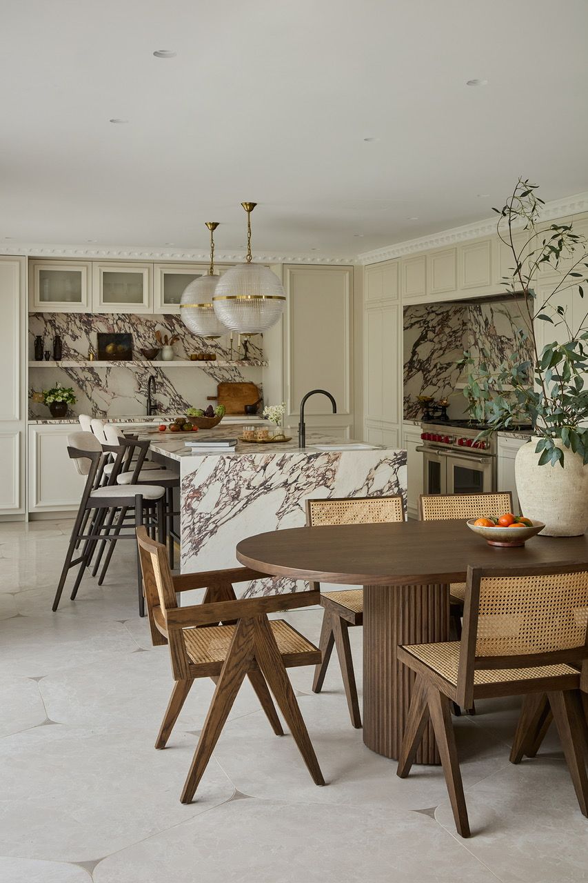 High-spec cream neutral kitchen at Love Locations featuring panelled walls, marble inlay, neutral tiled floor, modern cream upholstered bar stools, and a dark dining table with cane-backed chairs.