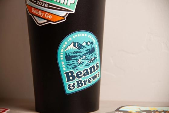 Close-up of a teal arched sticker reading "Now brewing in Spring Creek, Nevada" over a mountain and river illustration, applied to a matte black tumbler.