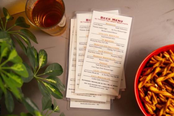 Overhead view of several laminated beer menus stacked and fanned beside a glass of amber beer, a plant and a bowl of pretzels.