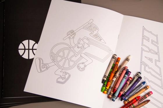 Coloring book opened to a line-art page of a basketball character holding a "Take Note" pennant, with crayons resting on the facing page of outlined block lettering.