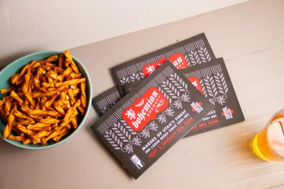 Overhead view of three Bohemian Brewery beer guides fanned on a table beside a bowl of pretzels and a glass of lager.