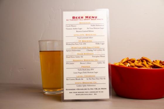 Laminated beer menu upright between a pint glass and a red bowl, headed "Beer Menu — $2 Tuesday all draft beer pints."