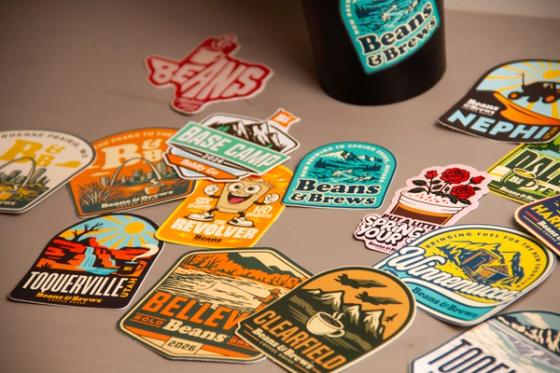Angled close view of overlapping die-cut stickers, including "The Revolver" cartoon coffee cup, a Texas-shaped "Beans" badge, and location designs for Toquerville, Belleville and Clearfield.