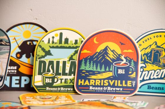 our location stickers standing upright, reading Nephi Utah, Dallas Texas, Harrisville UT and Winnemucca, each with an illustrated local scene and the Beans & Brews Coffee House logo.