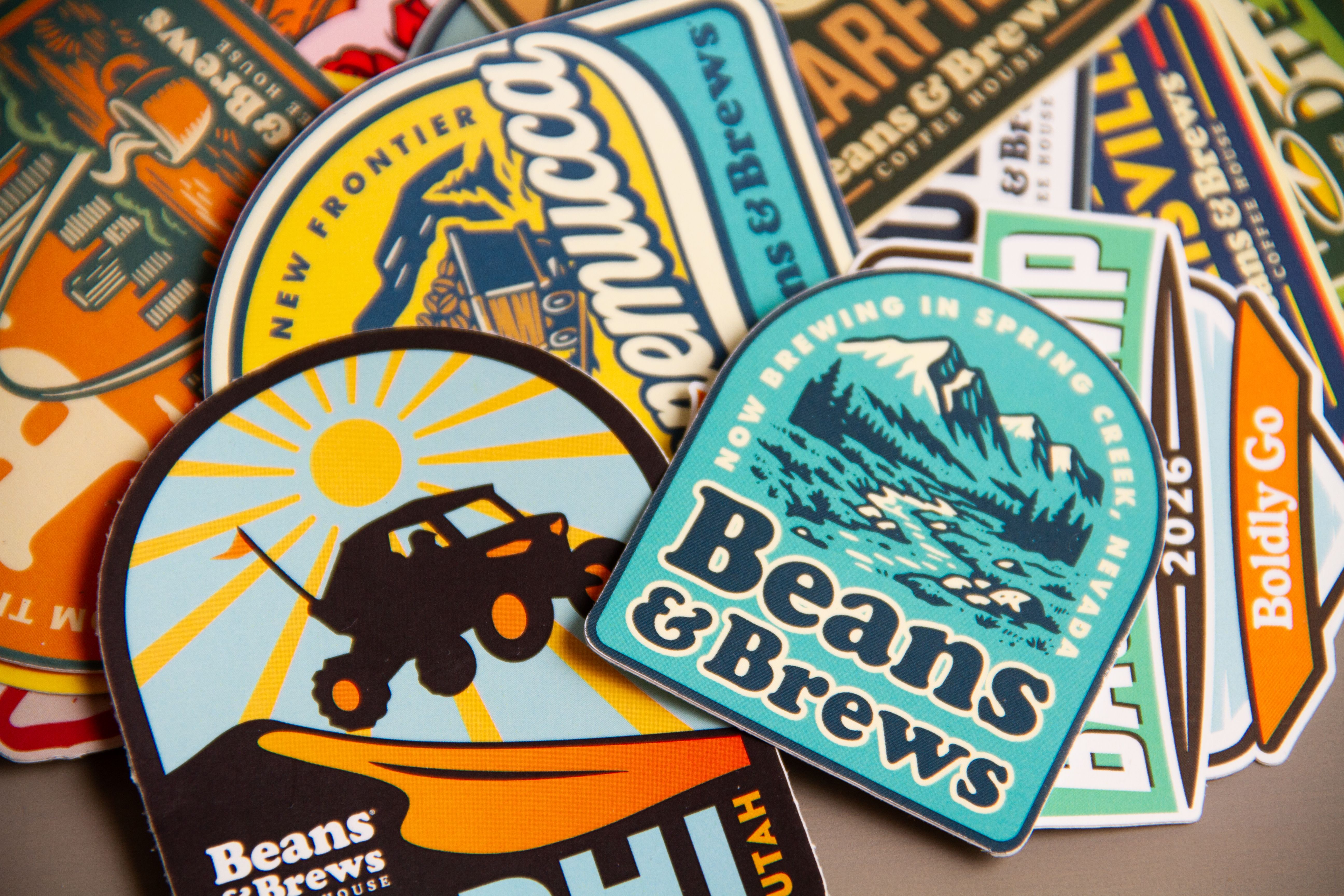 Tight overhead crop of stacked Beans & Brews stickers, with the Winnemucca, Spring Creek and Nephi designs in focus.