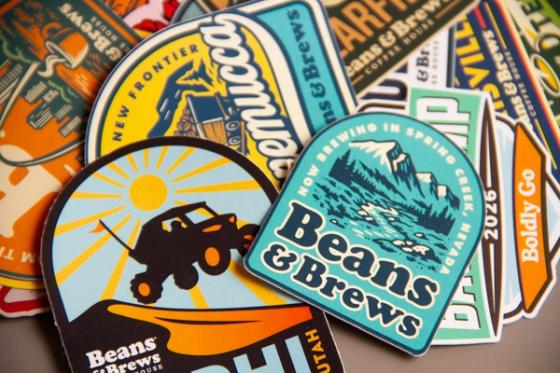 Tight overhead crop of stacked Beans & Brews stickers, with the Winnemucca, Spring Creek and Nephi designs in focus.