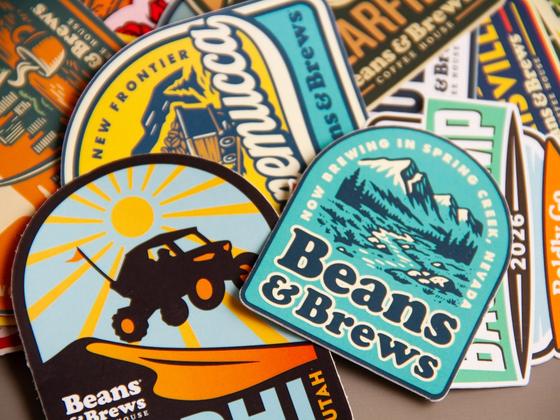 Tight overhead crop of stacked Beans & Brews stickers, with the Winnemucca, Spring Creek and Nephi designs in focus.