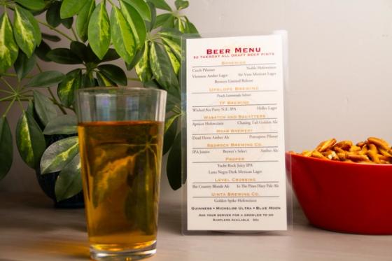 Laminated beer menu standing beside a pint of lager and a bowl of pretzels, listing draft offerings from Bohemian, TF Brewing, Wasatch and Squatters, Moab Brewery, RedRock, Proper, Level Crossing and Uinta.