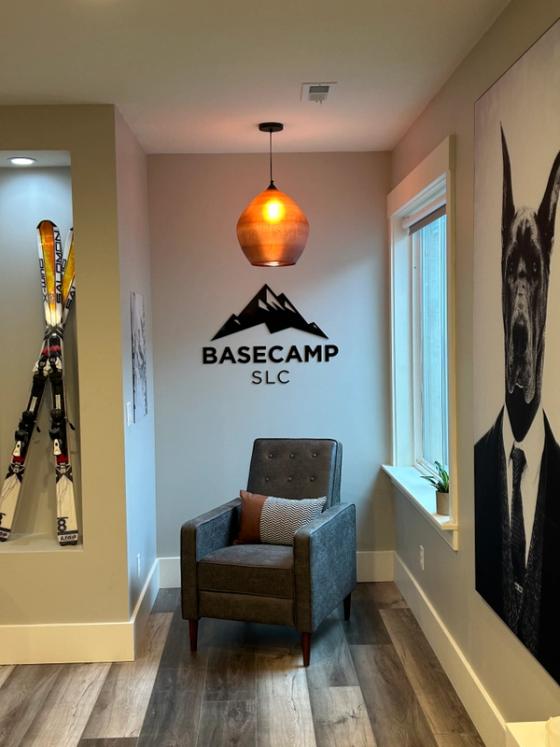 Dimensional black acrylic logo reading "BASECAMP SLC" with a mountain icon, mounted on a lobby wall above a chair.