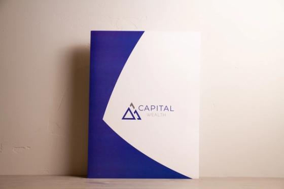 ront of the Capital Wealth presentation folder against a plain wall, showing the blue and white curve and centered logo.
