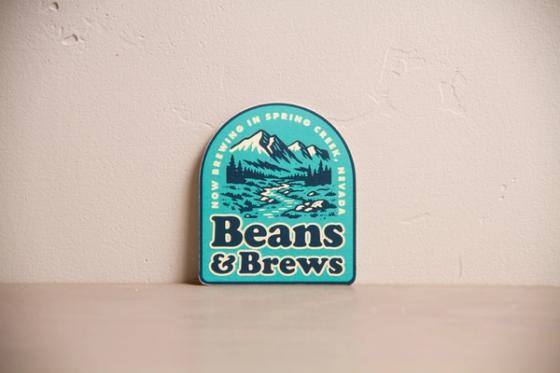 Single teal arched die-cut sticker reading "Now brewing in Spring Creek, Nevada" above the Beans & Brews wordmark.