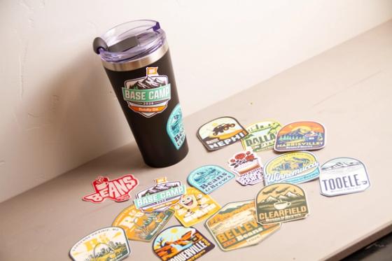 Branded tumbler standing on a ledge beside a spread of Beans & Brews location stickers for Nephi, Dallas, Harrisville, Winnemucca and Tooele.