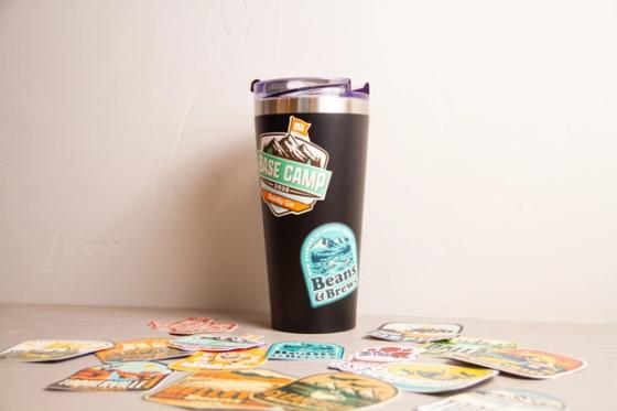 Black insulated tumbler with "Base Camp 2026 Boldly Go" and "Beans & Brews" stickers applied, with more location stickers scattered on the table in front.