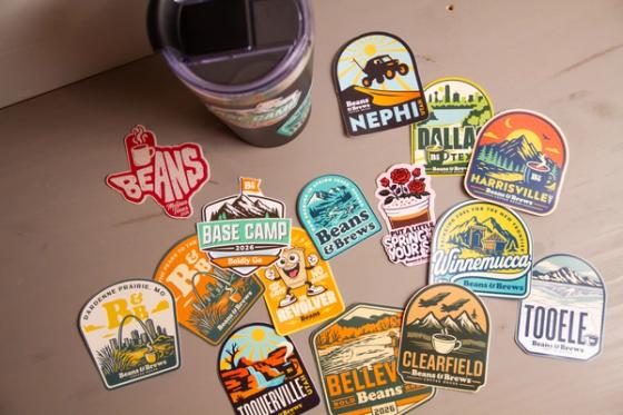 Overhead view of roughly fifteen Beans & Brews die-cut stickers fanned across a table, including location badges for Nephi, Dallas, Harrisville, Winnemucca, Tooele, Clearfield and Toquerville.