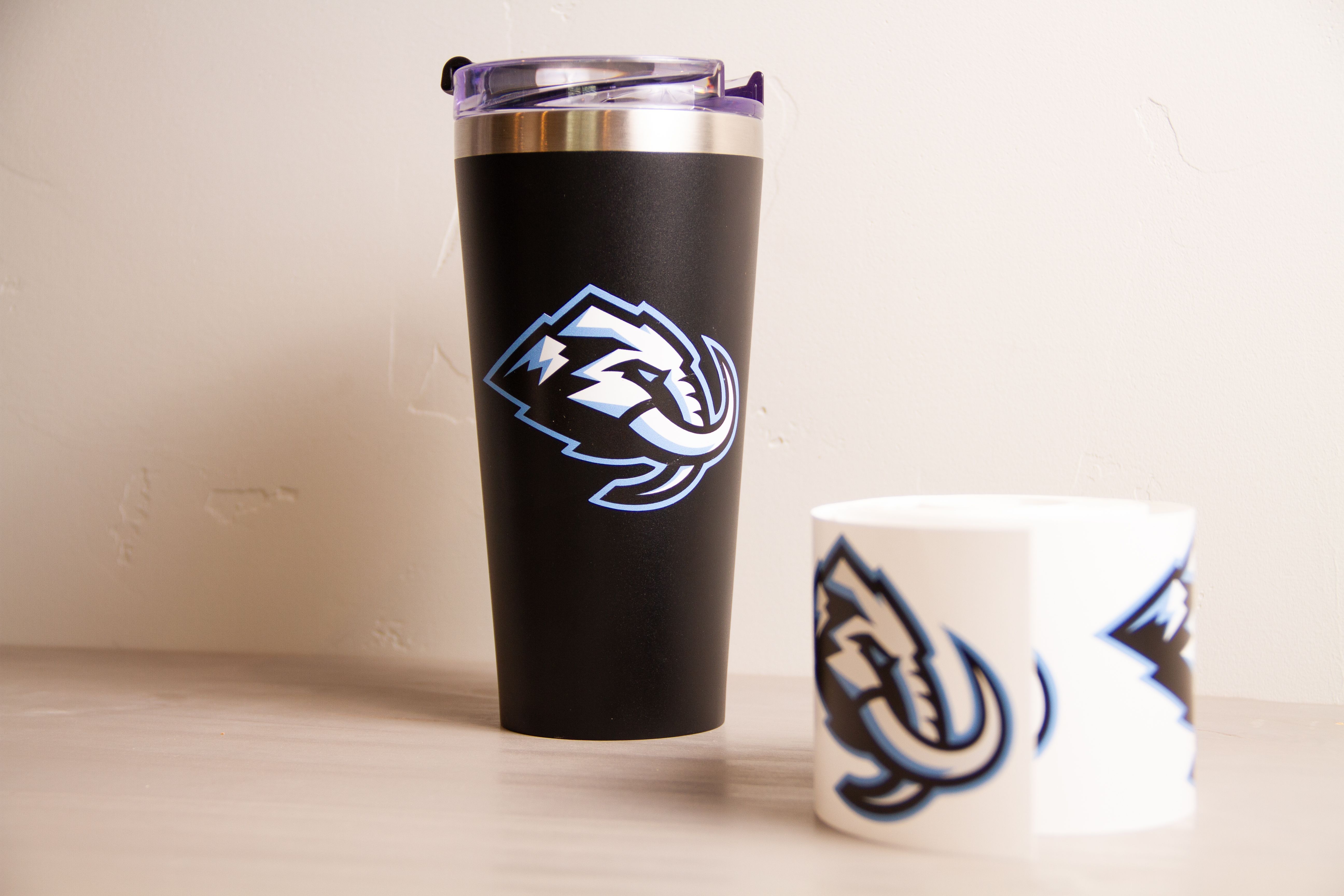 Black insulated tumbler with the Utah Mammoth logo applied, beside a roll of white vinyl printed with the same logo.