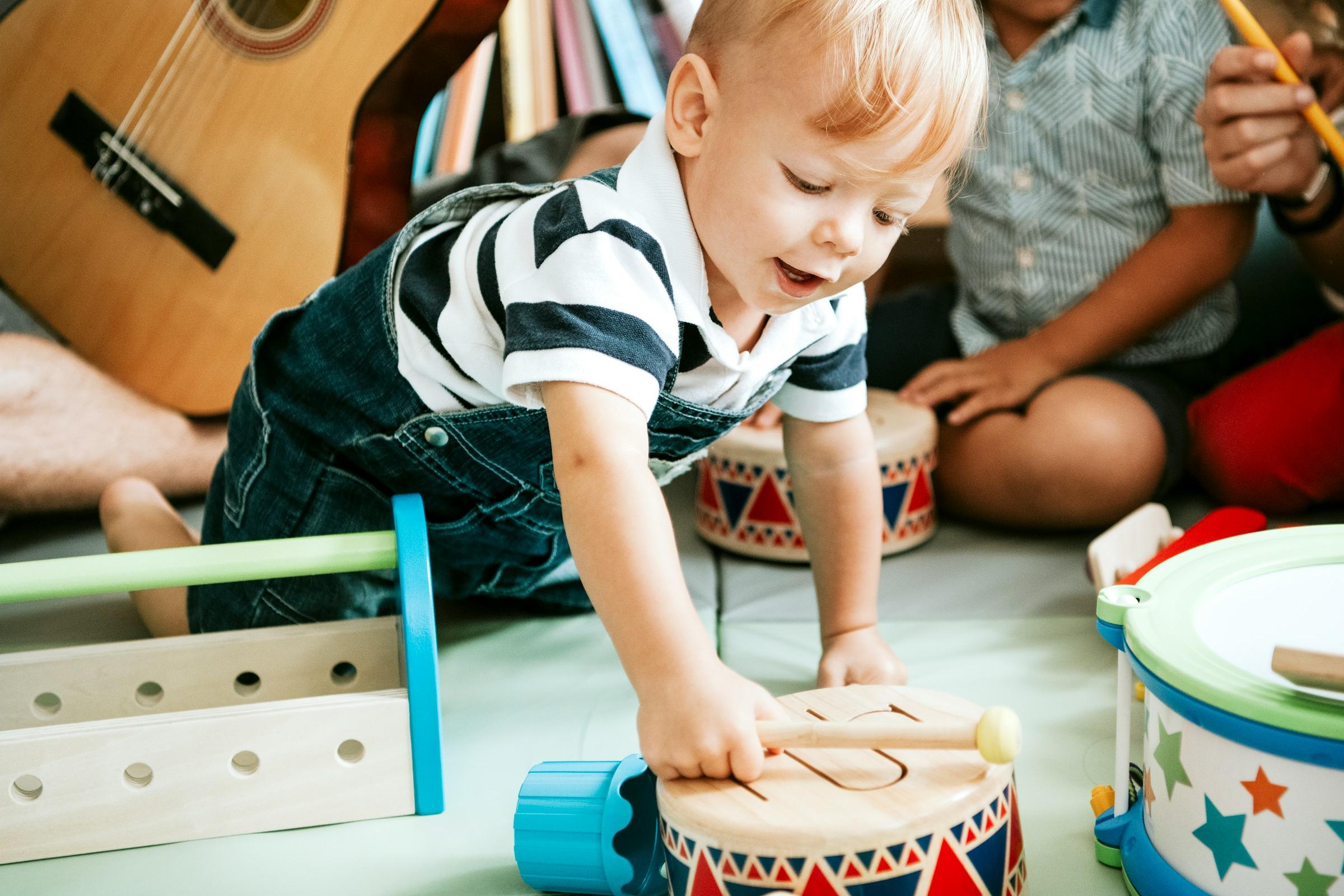 Toddler playing a wooden drum during a music session, surrounded by toy instruments and a guitar