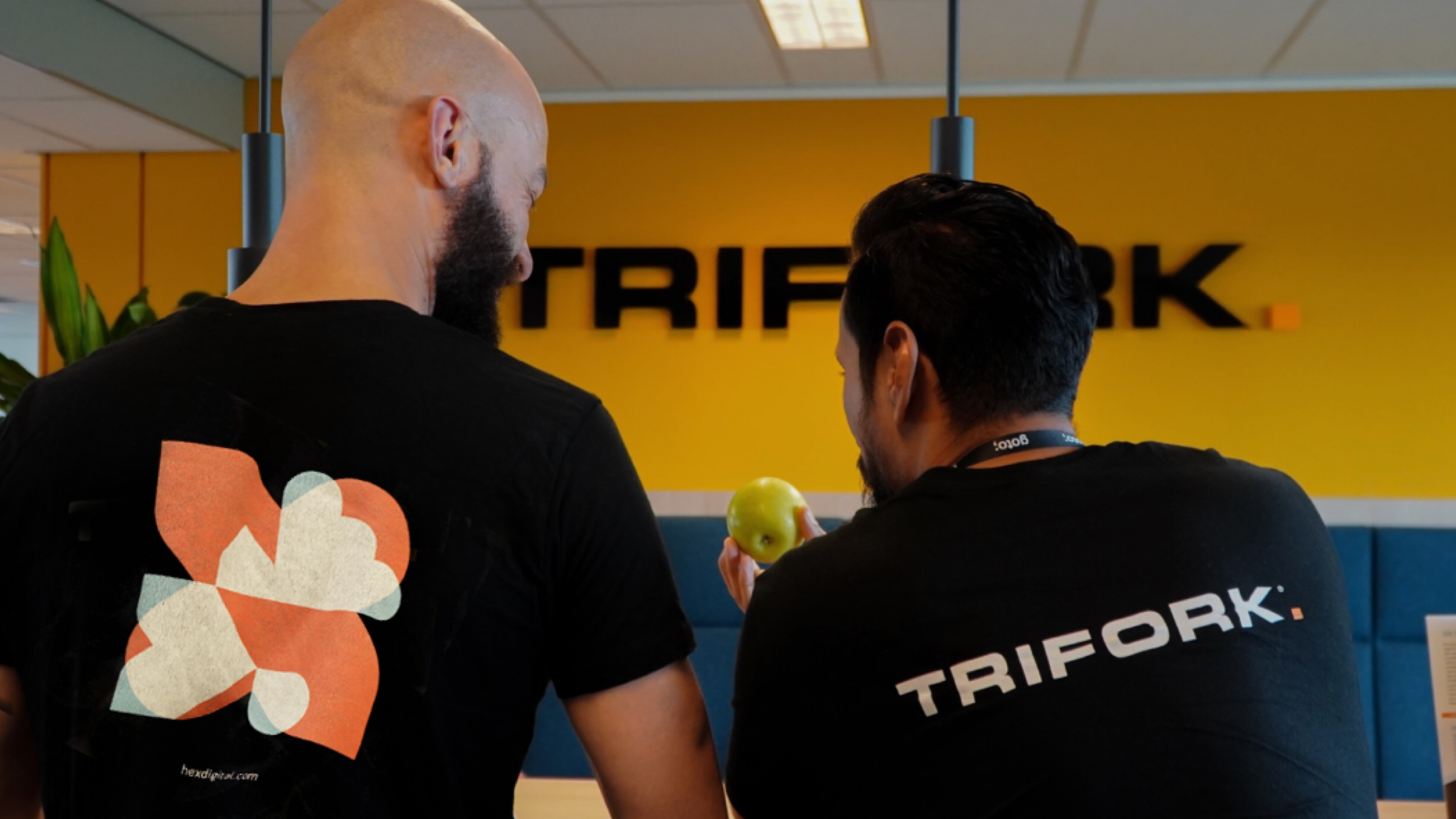 Two people seen from behind in front of a yellow Trifork wall sign, one in a Hex T-shirt and one in a Trifork T-shirt