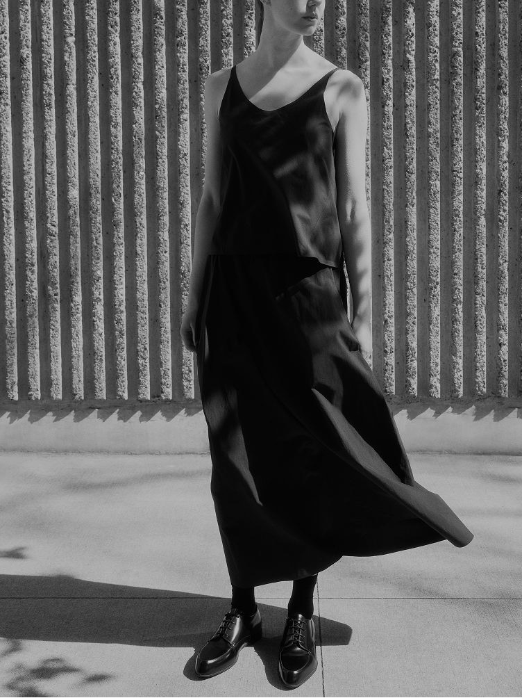 A person in a flowing dark camisole and skirt with pointed-toe oxford shoes stands against a textured wall in a black and white image.