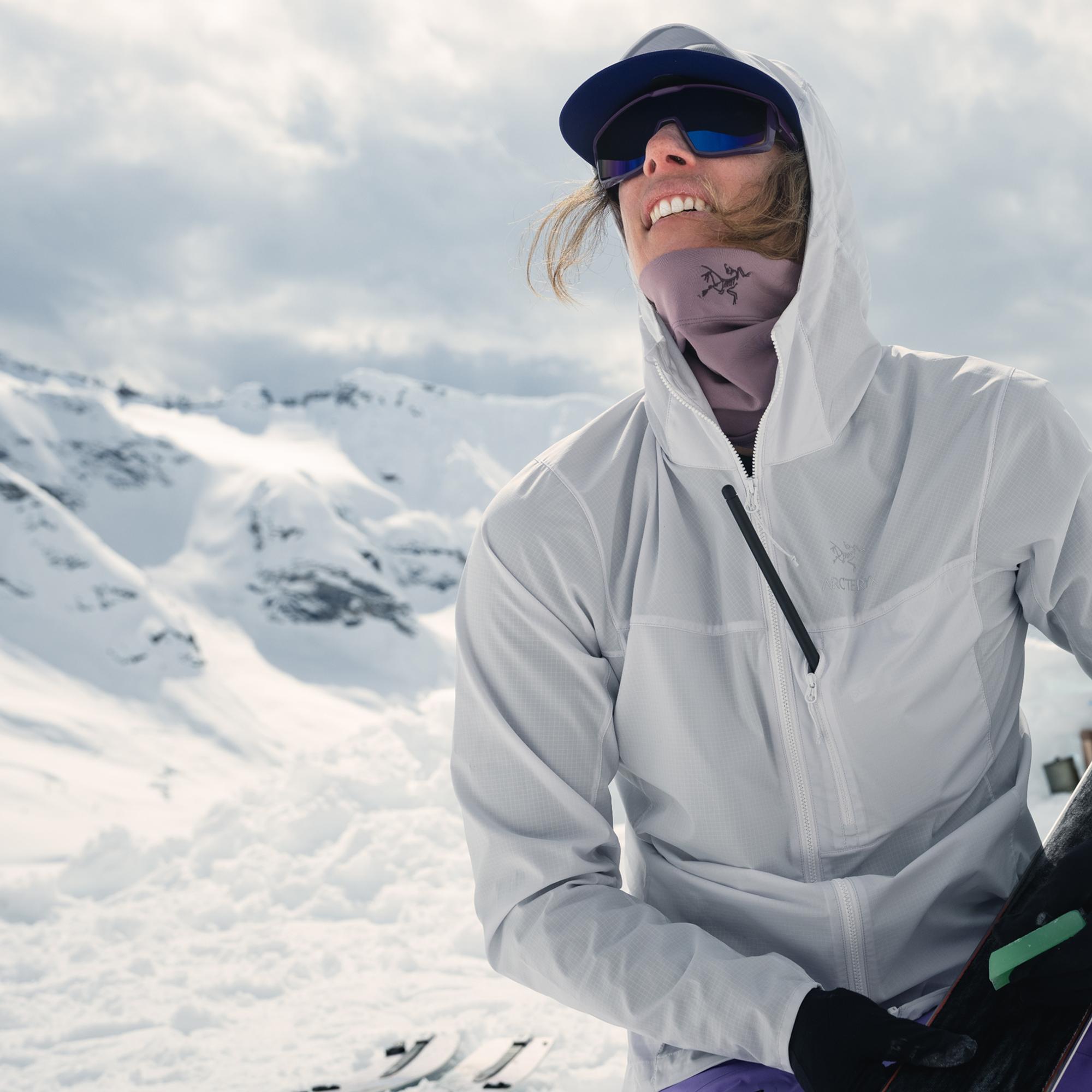 A person in a light jacket and sunglasses smiles, looking up at snowy mountains while holding skis.
