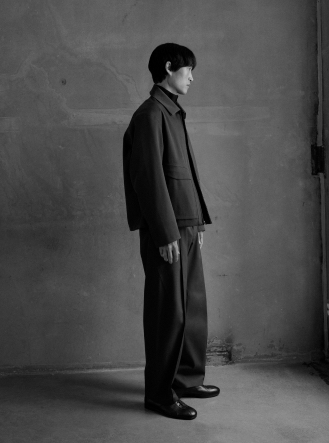 A man in a dark, minimalist outfit stands in profile against a textured wall, looking right, in a black and white photo.