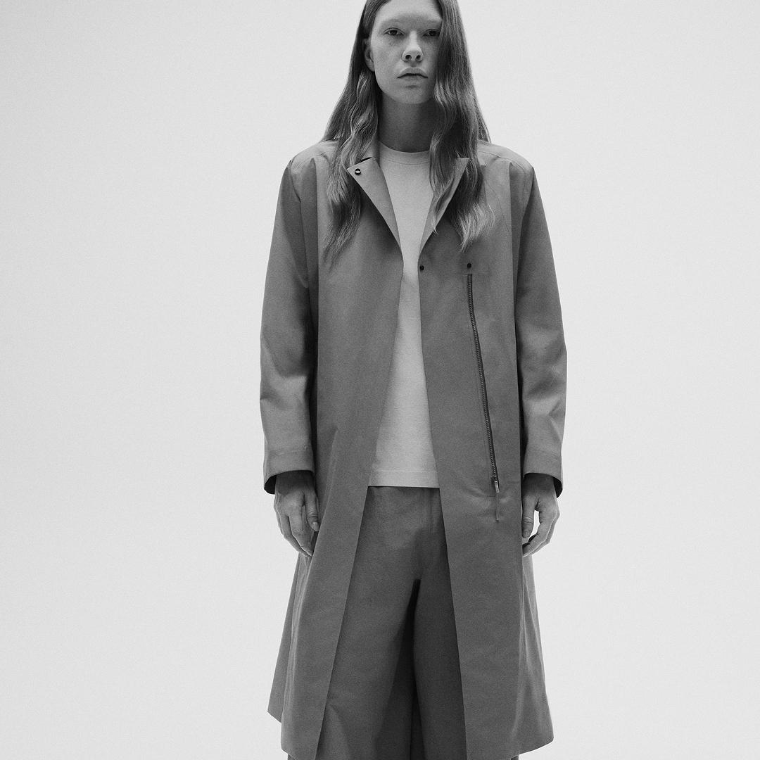 Black and white photo of a person with long hair wearing a minimalist long coat and pants.