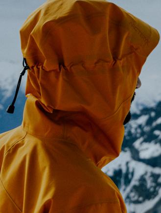 Back of a person wearing a yellow Arc'teryx hooded jacket against a snowy mountain backdrop.