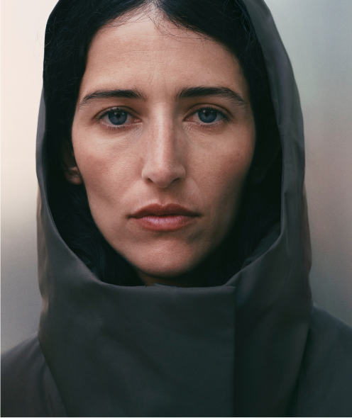 A woman with dark hair and blue eyes, wearing a dark hooded garment, looks directly at the camera with a neutral expression.