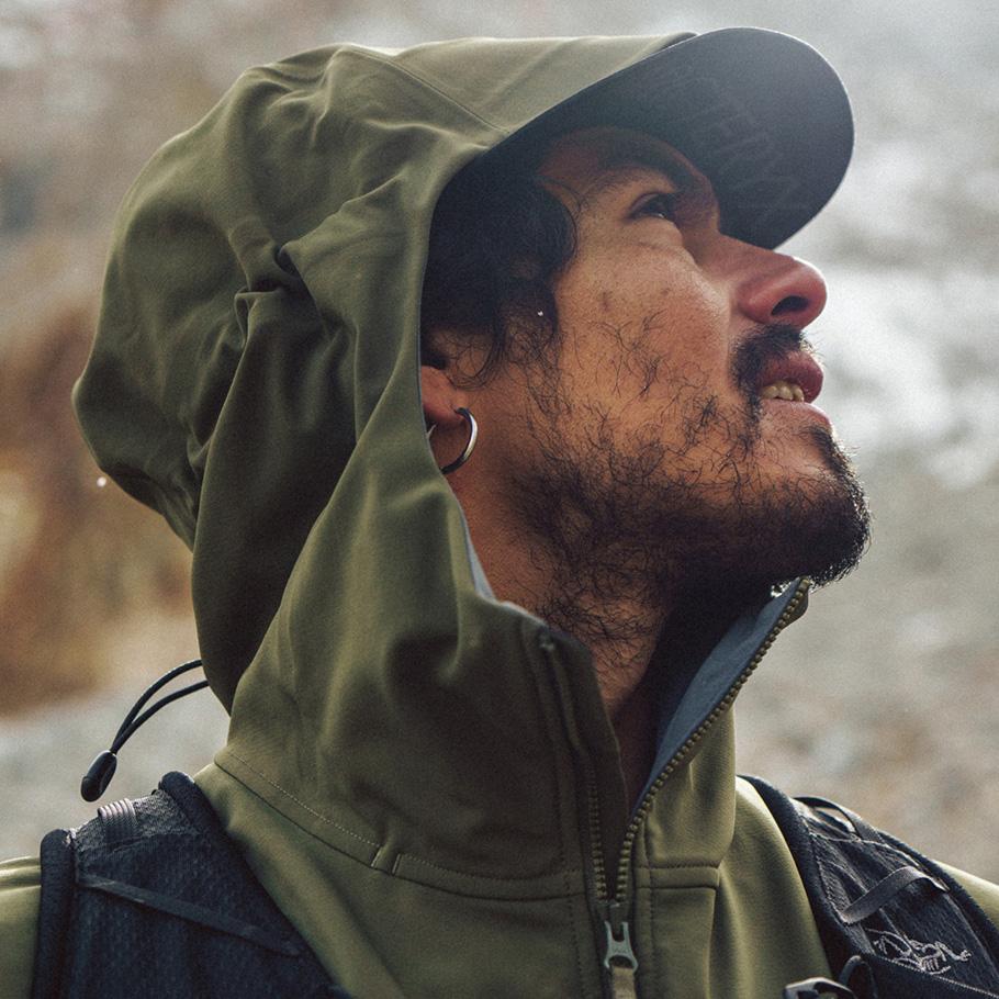 A man in an olive green hooded jacket with a cap looks up, smiling slightly.