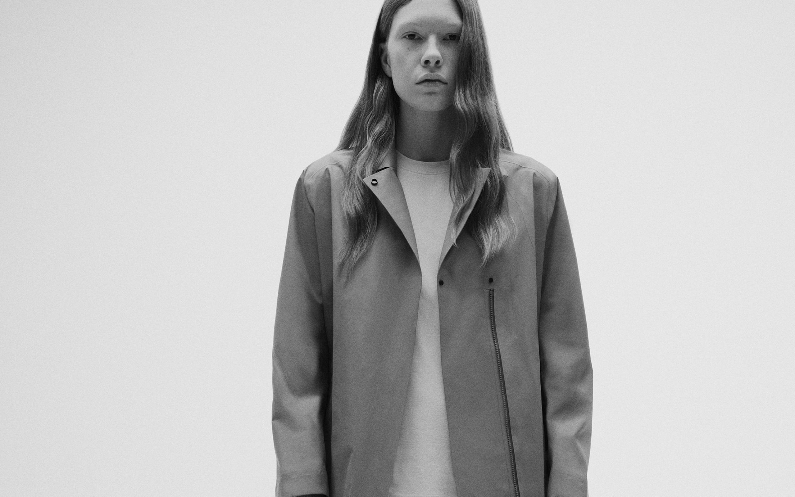 Black and white photo of a model with long, wavy hair wearing a minimalist jacket and t-shirt.