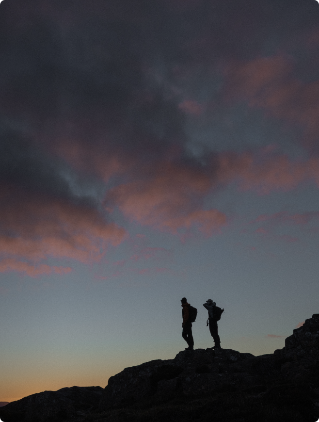 Two silhouetted hikers stand on a rocky ridge against a twilight sky with pink and grey clouds.