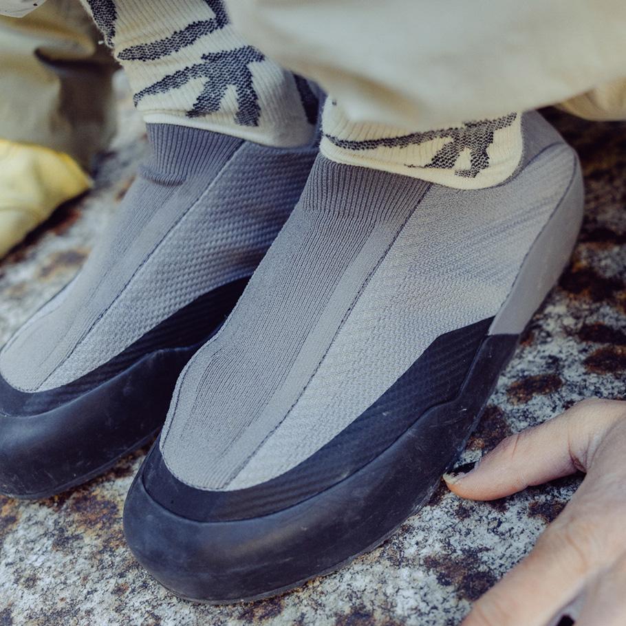 A pair of feet in grey slip-on shoes with black rubber soles and patterned socks stand on a rock surface.