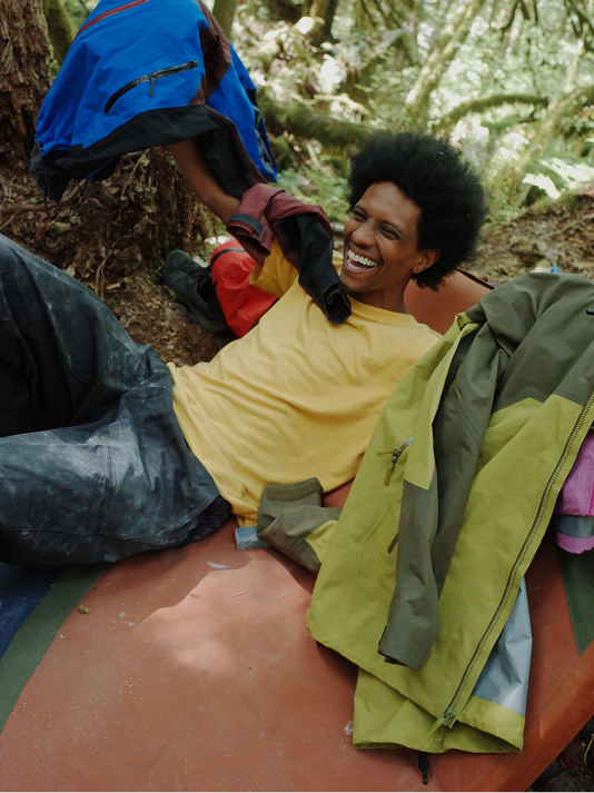 A man in a yellow shirt laughs heartily while relaxing among outdoor gear in a forest.