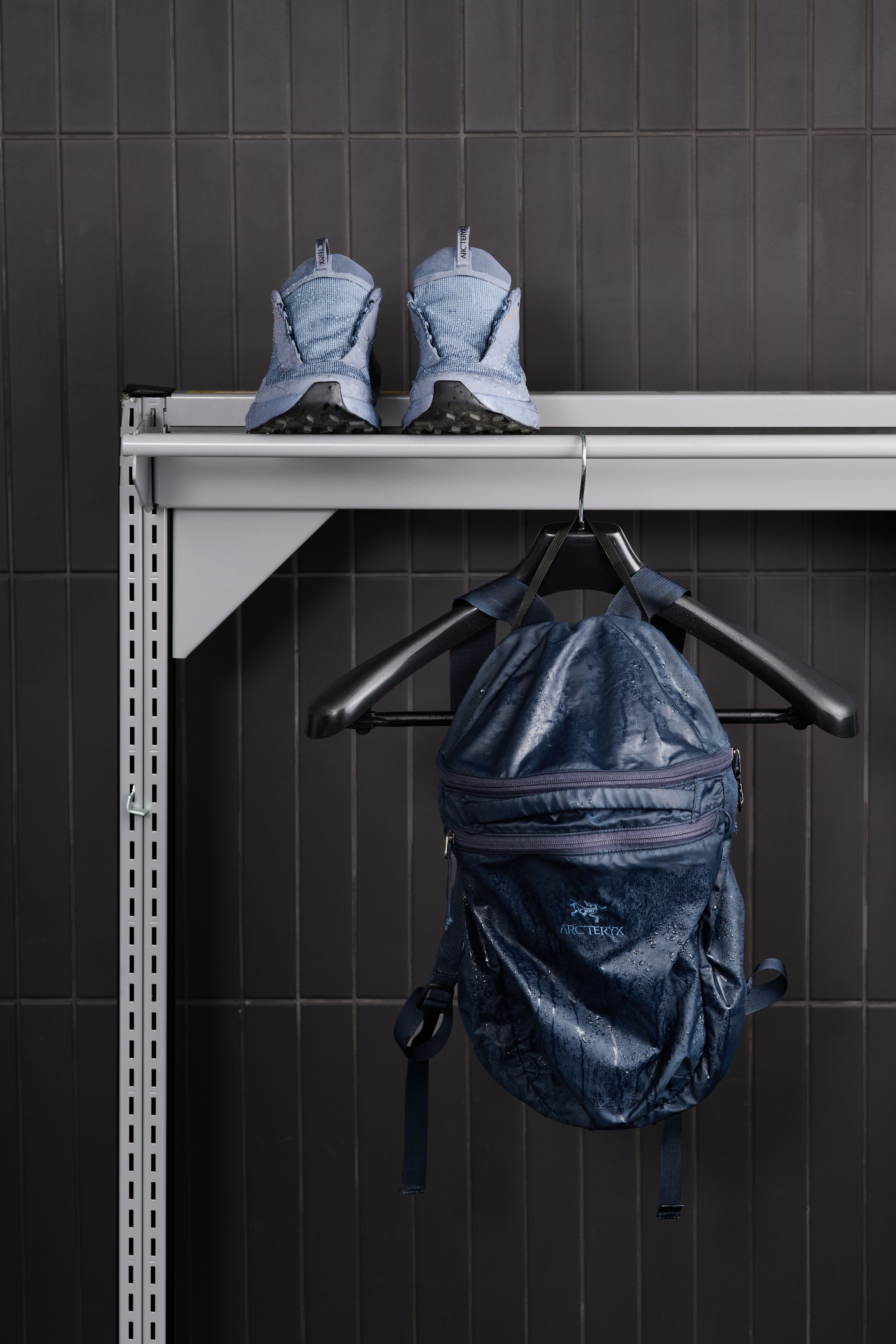 A pair of wet light blue sneakers on a shelf above a wet dark blue Arc'teryx backpack hanging on a hook, against a dark tiled wall.