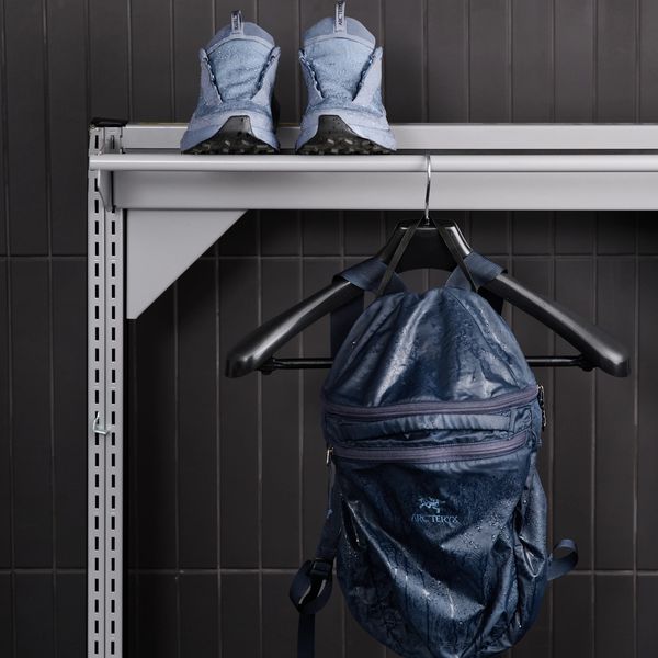 A pair of wet light blue sneakers on a shelf above a wet dark blue Arc'teryx backpack hanging on a hook, against a dark tiled wall.