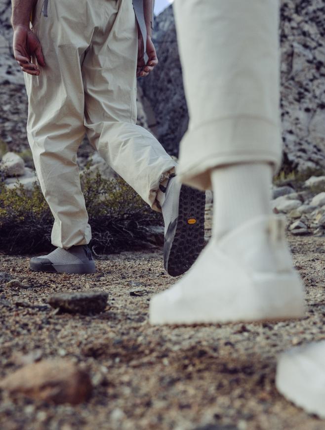 Two people wearing light outdoor pants and shoes walk on a rocky path; the foreground shoe has a dark textured sole with a yellow Vibram logo.